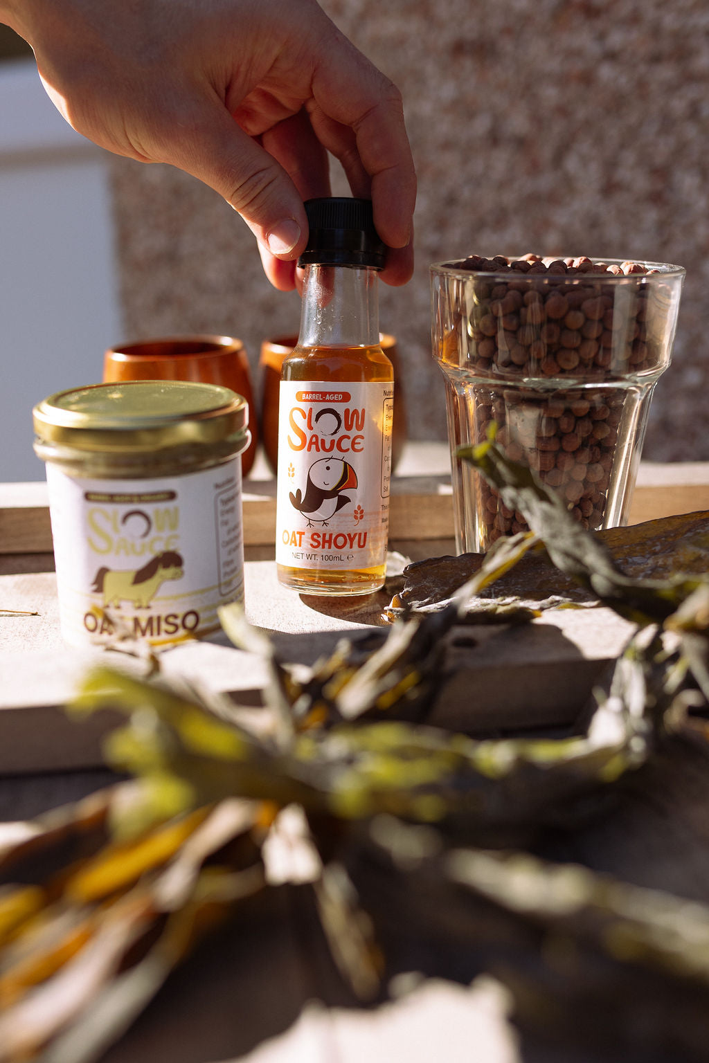 A hand picking up a bottle of Slow Sauce Oat Shoyu next to a jar of Oat Miso, with dried kelp and a jar of peas on a wooden table.